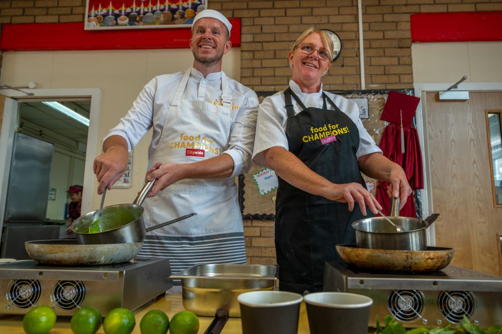 Two Development Chefs cooking at stovetops in a kitchen. The person on the left wears a white chef's uniform and hat, while the person on the right wears a black apron over a white shirt. The aprons read 'food for CHAMPIONS'. On the counter are limes and two cups.