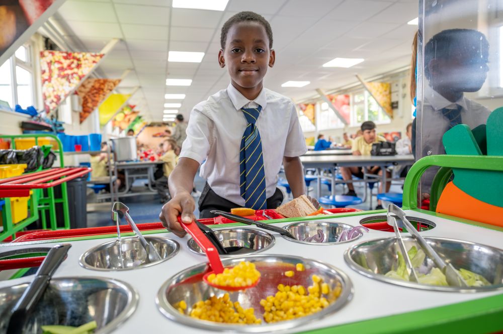 A child in a school cafeteria serves food from a salad bar, wearing a white shirt and blue striped tie. The child's face is blurred for privacy. The salad bar includes corn, lettuce, and other vegetables in metal containers with serving utensils. In the background, other children are seated at tables.
